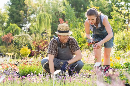 A man and a girl working together in a lush garden, with the man crouching near a flower bed containing a variety of small, colorful flowering plants and shrubs. The garden features a neatly maintained lawn area with dense, evenly cut grass in the foreground. Behind them, there are tall trees with green foliage, a border of bushes and flowering plants, and a mixture of natural tones ranging from vibrant greens to soft pinks and purples. The man is wearing a straw hat, a checked shirt, and gardening gloves, while the girl is dressed in a sleeveless top and denim shorts, holding garden tools and leaning slightly forward as they tend to the plants. The sunlight filters through the trees, illuminating the outdoor space with bright, natural light, creating a warm and inviting atmosphere characteristic of a well-maintained landscaped garden suitable for local gardening services in Peckham or nearby areas. This scene indicates active gardening or planting work, emphasizing the care of flower beds and outdoor landscape features, reflecting professional landscaping and gardening efforts.