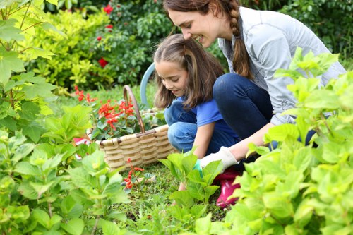 A woman and two young girls are gardening outdoors in a lush, green garden. The woman, with long brown hair in braids, is crouched down and smiling as she supervises the girls, who are also engaged in planting or tending to garden plants. The area features a dense collection of vibrant green foliage, including large leafy plants and flowering shrubs with red blossoms. In the background, there are more garden plants and a mixture of hedges and trees creating a natural, landscaped outdoor space. The ground is covered with well-maintained grass, and small patches of soil are visible where the children are working. The scene is brightly lit, suggesting a sunny day with clear weather. The image reflects a peaceful urban garden environment, indicative of a backyard or front garden space in Peckham, and subtly aligns with gardening and outdoor maintenance services offered by Landscapers Peckham.