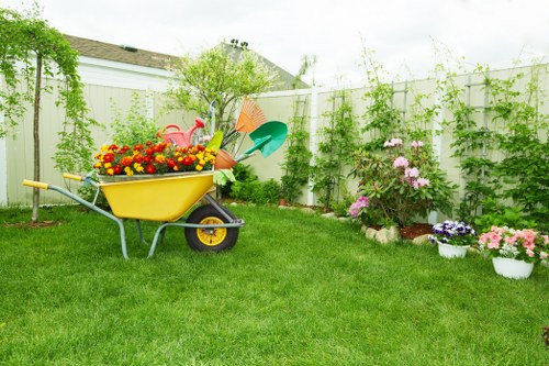 A well-maintained backyard garden featuring a lush, green lawn with evenly cut grass in the foreground. In the middle of the yard, there is a yellow wheelbarrow filled with vibrant gardening supplies, including brightly coloured flowers and decorative garden ornaments, such as a pink flamingo. The garden is bordered by a wooden fence painted white, which is partially covered by climbing plants and greenery. To the right, there are flower beds containing blooming shrubs and a variety of flowering plants in shades of pink, purple, and white, with some potted flowers placed along the border. Behind the fence, a few tall trees and additional foliage provide a natural backdrop, contributing to the outdoor setting's privacy. The scene is illuminated by natural daylight under a mostly cloudy sky, creating a soft, even light across the scene. The setting suggests an outdoor space cared for by professional gardening services, with a focus on lawn care, planting, and overall garden maintenance, ideal for a residential area in Peckham or nearby southeast London suburbs. Landscaping and gardening elements are clearly visible, supporting local gardening keywords naturally.