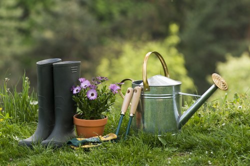 The image depicts a well-maintained garden scene with a focus on gardening tools and plant care items placed on lush, green grass. In the foreground, there are a pair of black rubber gardening boots standing upright, alongside a small terracotta pot filled with vibrant purple flowers, which are in full bloom. Next to these, a set of small pruning shears and hand tools with beige and green handles are laid out, ready for garden maintenance tasks. A shiny, metal watering can with a curved handle and a long spout is positioned nearby, suggesting ongoing or upcoming watering activity. Behind the tools, there is a background of soft greenery with blurred trees and bushes, indicating a natural outdoor environment, possibly part of a backyard or front garden in Peckham. The scene is lit by natural daylight, with a slightly overcast sky providing diffuse lighting that highlights the rich textures of the soil, plant leaves, and Garden tools. This setup exemplifies typical outdoor garden maintenance, with clear connections to professional gardening and landscaping services offered by Landscapers Peckham around the local area, including the postcode in SE15. The garden appears tidy and inviting, reflecting attentive care suitable for clients seeking planting, lawn care, and outdoor beautification services in London.