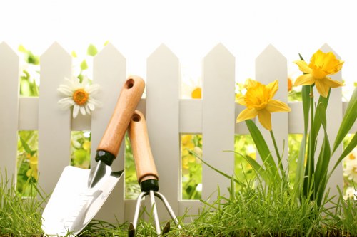 A close-up view of a small garden area in Peckham, showing a white picket fence partially obscured by vibrant yellow daffodils and white daisies growing along the edge of a well-maintained lawn with lush, green grass. In front of the fence, two gardening tools—a trowel and a hand fork with wooden handles—are planted in the soil, suggesting ongoing gardening activities. The environment appears bright and sunny, with natural light illuminating the foliage and flowers, creating a cheerful outdoor scene suitable for professional gardening and landscaping maintenance by Landscapers Peckham.