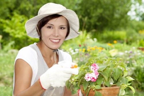 A woman wearing a wide-brimmed hat, white gardening gloves, and a white short-sleeved shirt is smiling while tending to a potted flowering plant with pink blossoms. She is in an outdoor garden setting with a well-maintained lawn, green foliage, and a variety of flowering plants visible in the background. The garden features a mix of grassy areas, flower beds, and shrubs, suggesting regular outdoor maintenance and gardening activities. The natural daylight indicates a bright, clear day, emphasizing a healthy and vibrant environment. This scene demonstrates typical garden care actions, relevant to landscaping and gardening services offered by companies like Landscapers Peckham, and showcases a cultivated outdoor space within urban or suburban surroundings, possibly in or near Peckham or South London.