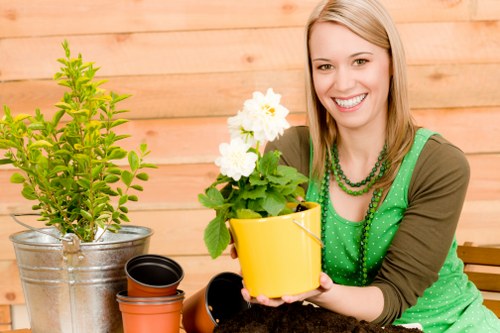 The image shows a woman with light brown hair and a bright smile, sitting outdoors in a garden setting. She is wearing a green polka-dot top with a brown jacket and a matching green beaded necklace. In front of her, she holds a yellow plant pot containing a healthy, blooming white flower plant. To her left, there is a larger galvanized metal container housing a lush green shrub with vibrant, dense foliage. On the table in front of her, there are several small black and terracotta-colored plant pots, some empty and others partially filled with soil, indicating gardening activity. The background features a wooden garden fence or paneling, suggesting a private backyard or landscaped outdoor space. The setting appears bright with natural daylight, indicating a sunny day, ideal for gardening. The overall scene provides a natural connection to gardening and outdoor maintenance services, illustrating a well-maintained, inviting garden environment that could benefit from professional landscaping or planting services offered by companies like Landscapers Peckham.