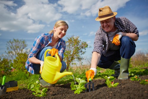 A young woman and an older man are working together in a garden, planting or tending to small leafy green plants in rich, dark soil. The woman is wearing a blue checked shirt and yellow gardening gloves, holding a yellow watering can with a black spout, while the man is dressed in a straw hat, plaid shirt, and green rubber boots, also wearing orange gloves. They are both smiling and focused on their gardening tasks, indicating a friendly and collaborative outdoor activity. The background features a clear blue sky with some scattered clouds, along with a distant hedge or row of trees, suggesting a well-maintained garden or landscaped outdoor space typical of residential properties in Peckham. The garden bed appears to have neat, defined edges, with a mix of soil and young plants that are evenly spaced, and the overall setting conveys a peaceful, productive environment suitable for gardening and landscape maintenance services provided by Landscapers Peckham.
