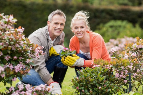 A man and a woman are kneeling in a well-maintained garden with a lush, green lawn and colorful flowering shrubs. They are smiling and wearing gardening gloves, actively tending to the plants, with the man holding a small potted plant and the woman supporting it. In the background, there are various green trees and hedges, suggesting a spacious outdoor residential garden in Peckham, south London. The scene is lit with natural daylight, possibly on a clear, slightly cloudy day, highlighting the vibrant colors of the flowers and the healthy appearance of the foliage. The garden surface features both soil beds and neatly trimmed grass, with some paving stones visible along the edge of the planting area, indicating a structured and manicured outdoor space suitable for landscaping and gardening services by Landscapers Peckham.